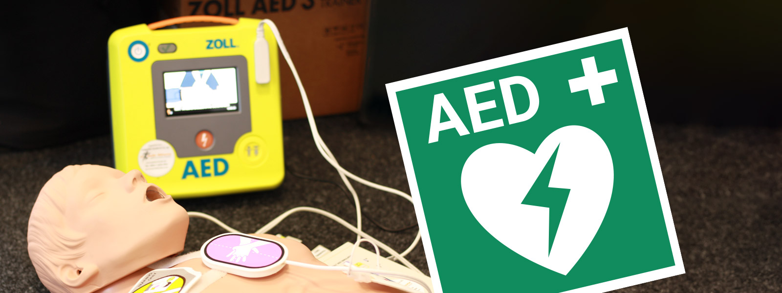 Hinweisschild AED Defibrillator, links eine Demonstationspuppe mit angelegten Defi-Pads